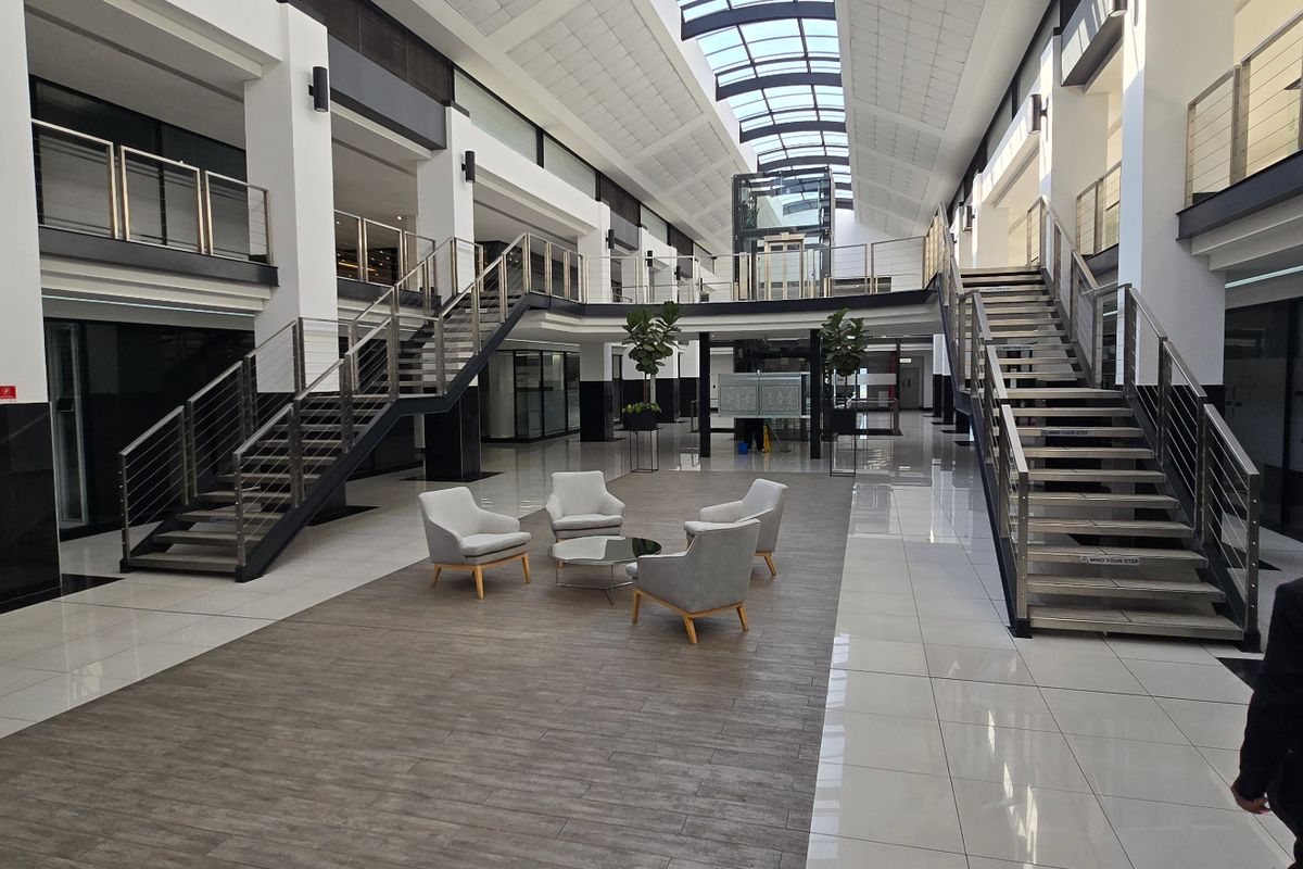 Spacious commercial atrium featuring a central skylight, glass elevator, and dual staircases with modern stainless steel railings and polished tile flooring.