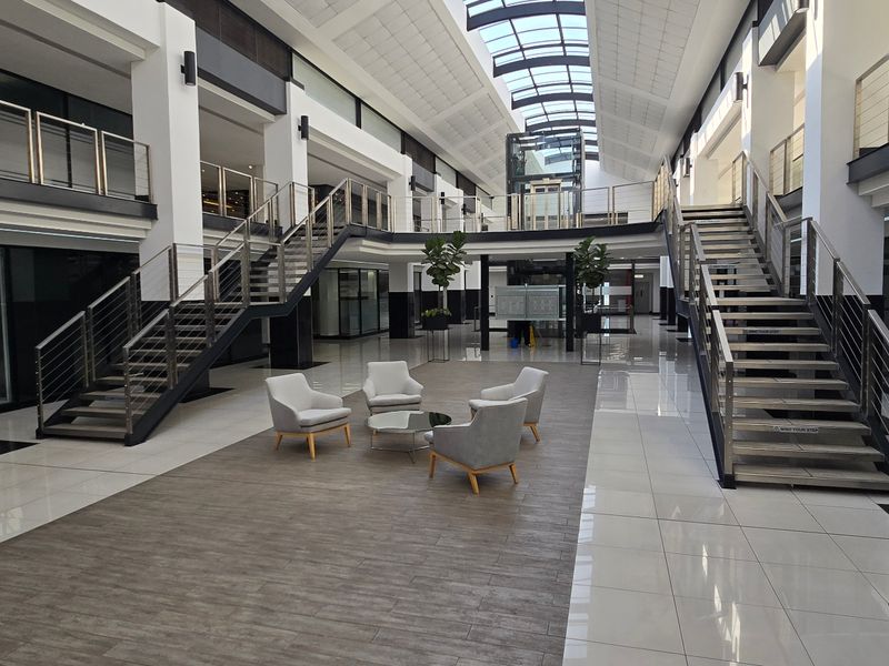 Spacious commercial atrium featuring a central skylight, glass elevator, and dual staircases with modern stainless steel railings and polished tile flooring.
