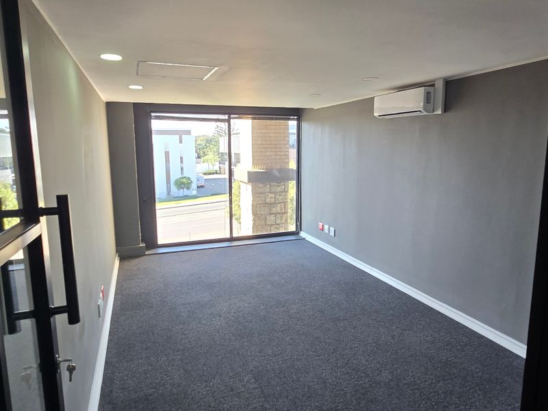 A modern, empty office space featuring grey carpeting, recessed lighting, a wall-mounted air conditioning unit, and black-framed glass partitions.