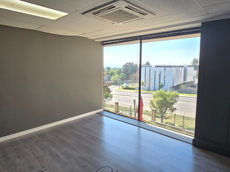 A bright, empty office space featuring modern laminate flooring, a suspended ceiling with integrated air conditioning, and large windows offering an exterior view.