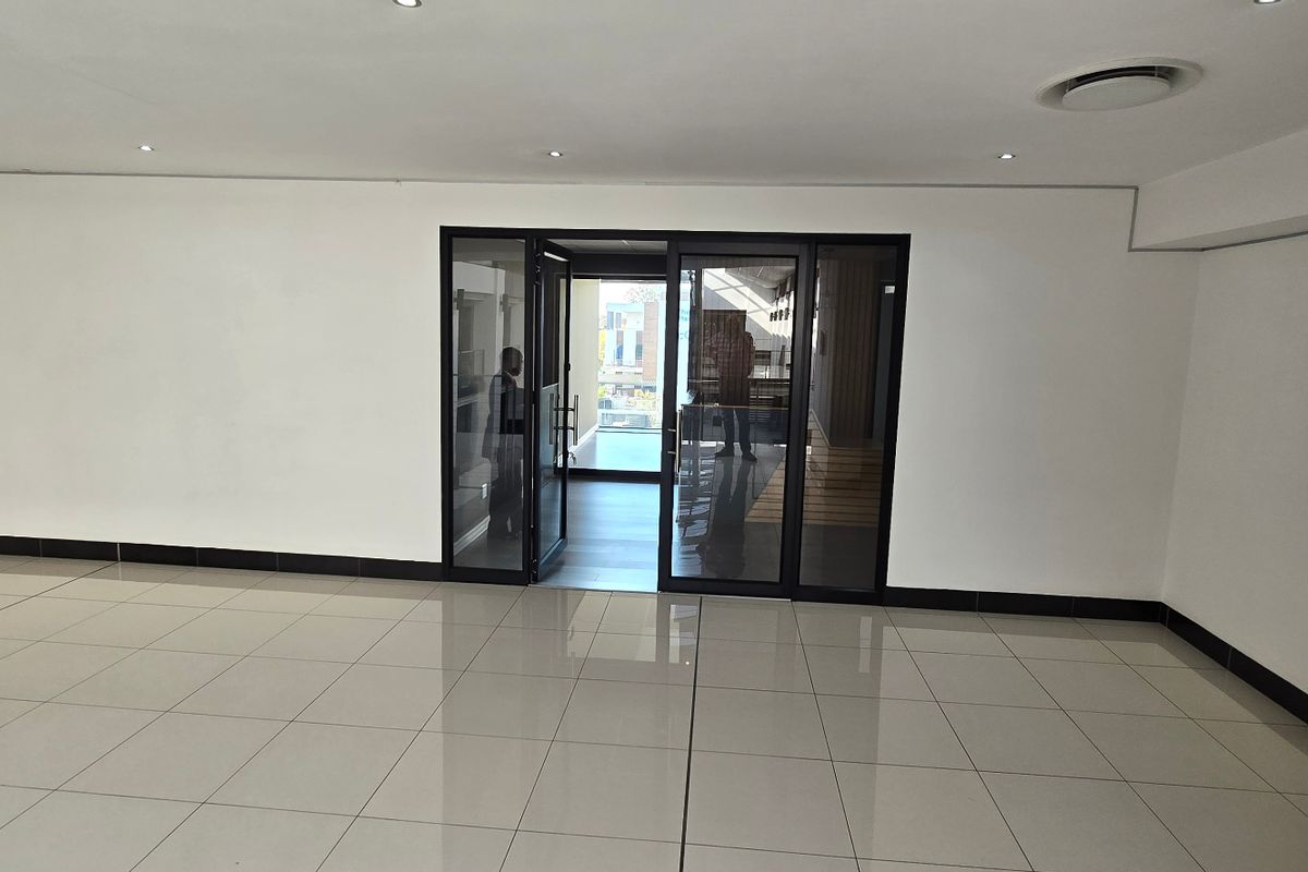 A spacious, empty commercial office area featuring modern porcelain floor tiles, recessed downlighting, and black aluminium framed glass entrance doors.
