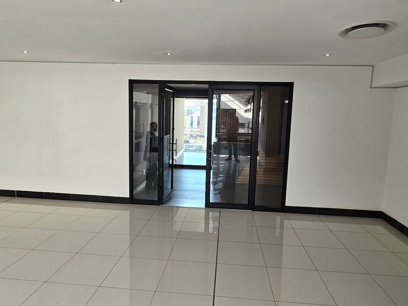 A spacious, empty commercial office area featuring modern porcelain floor tiles, recessed downlighting, and black aluminium framed glass entrance doors.