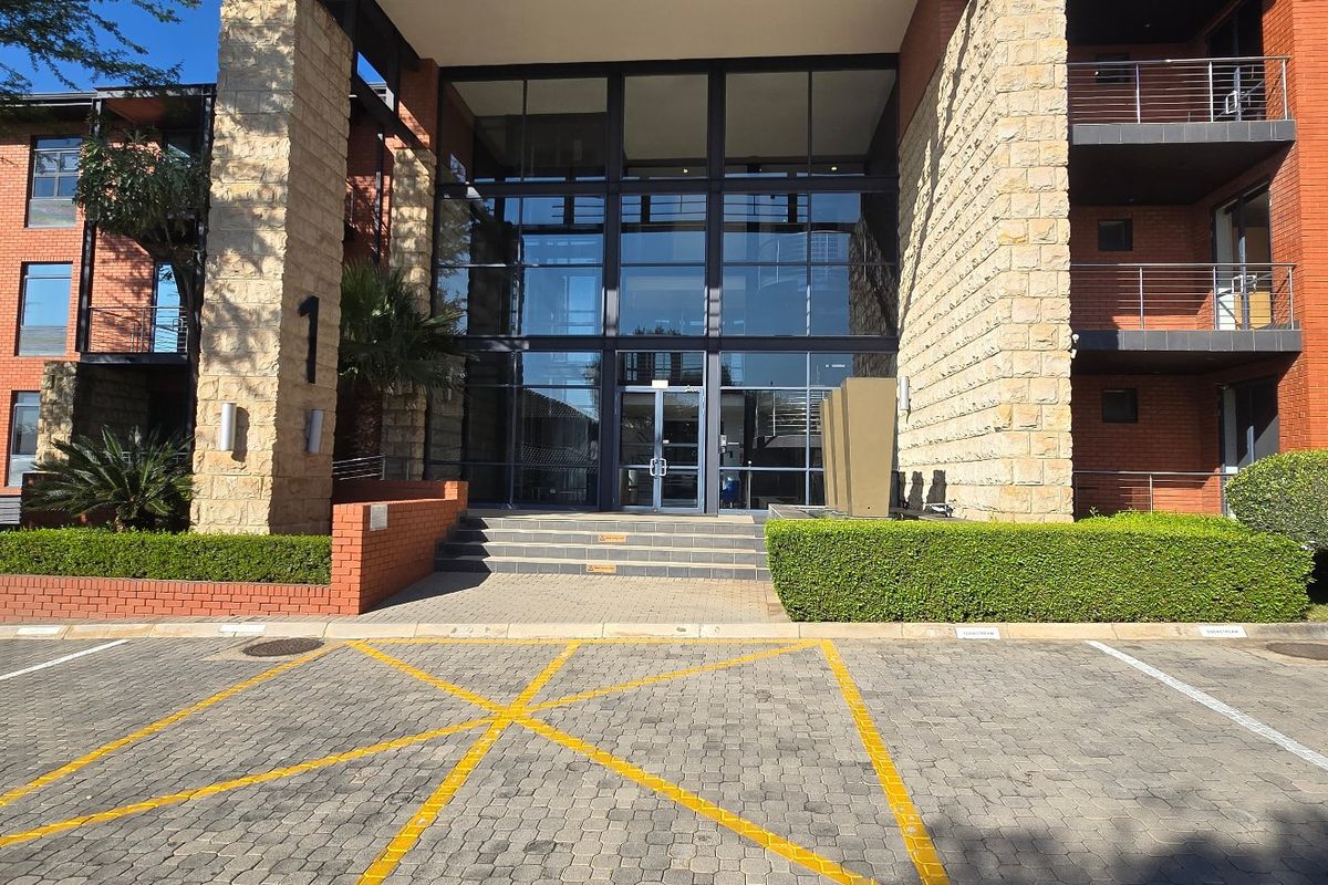 Modern commercial building entrance featuring a prominent glass facade, stone and brick exterior finishes, and a paved parking area with landscaped surroundings.