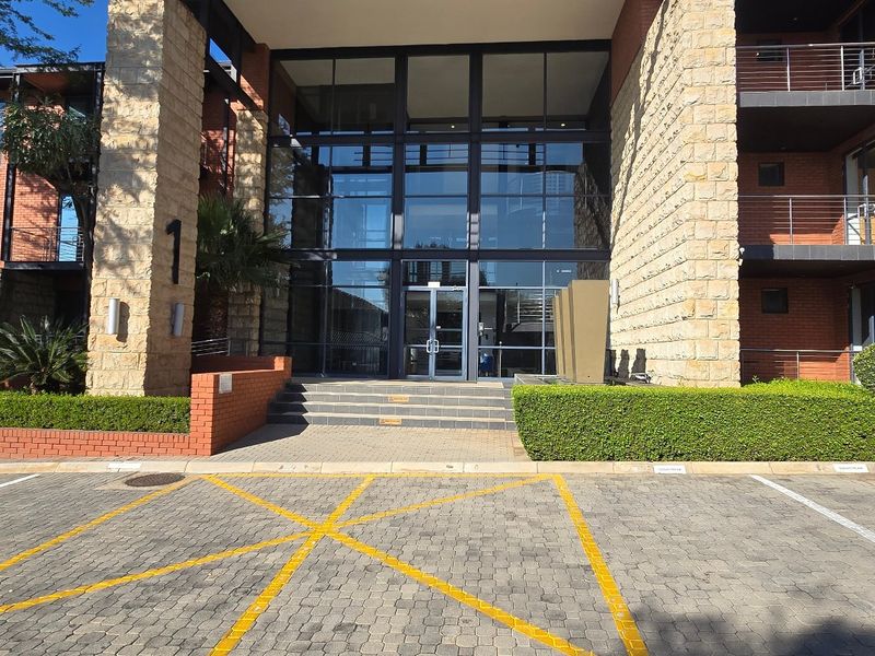 Modern commercial building entrance featuring a prominent glass facade, stone and brick exterior finishes, and a paved parking area with landscaped surroundings.