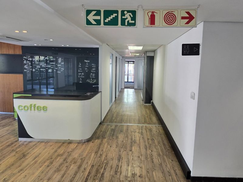 A bright commercial hallway featuring wood-look laminate flooring, a reception counter, and clear emergency exit signage leading to office spaces.