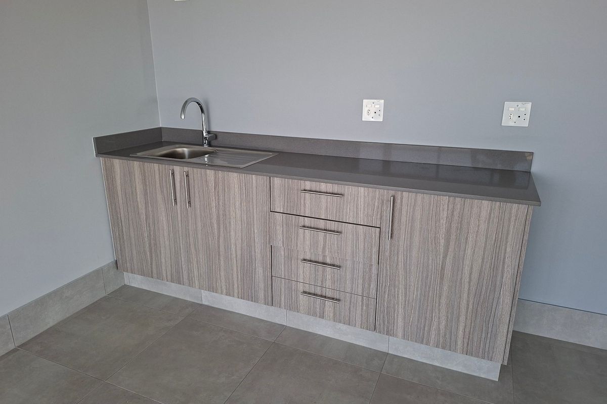 A clean, modern kitchenette featuring wood-grain cabinetry, a stone countertop, and a stainless steel sink, set against a neutral grey wall.