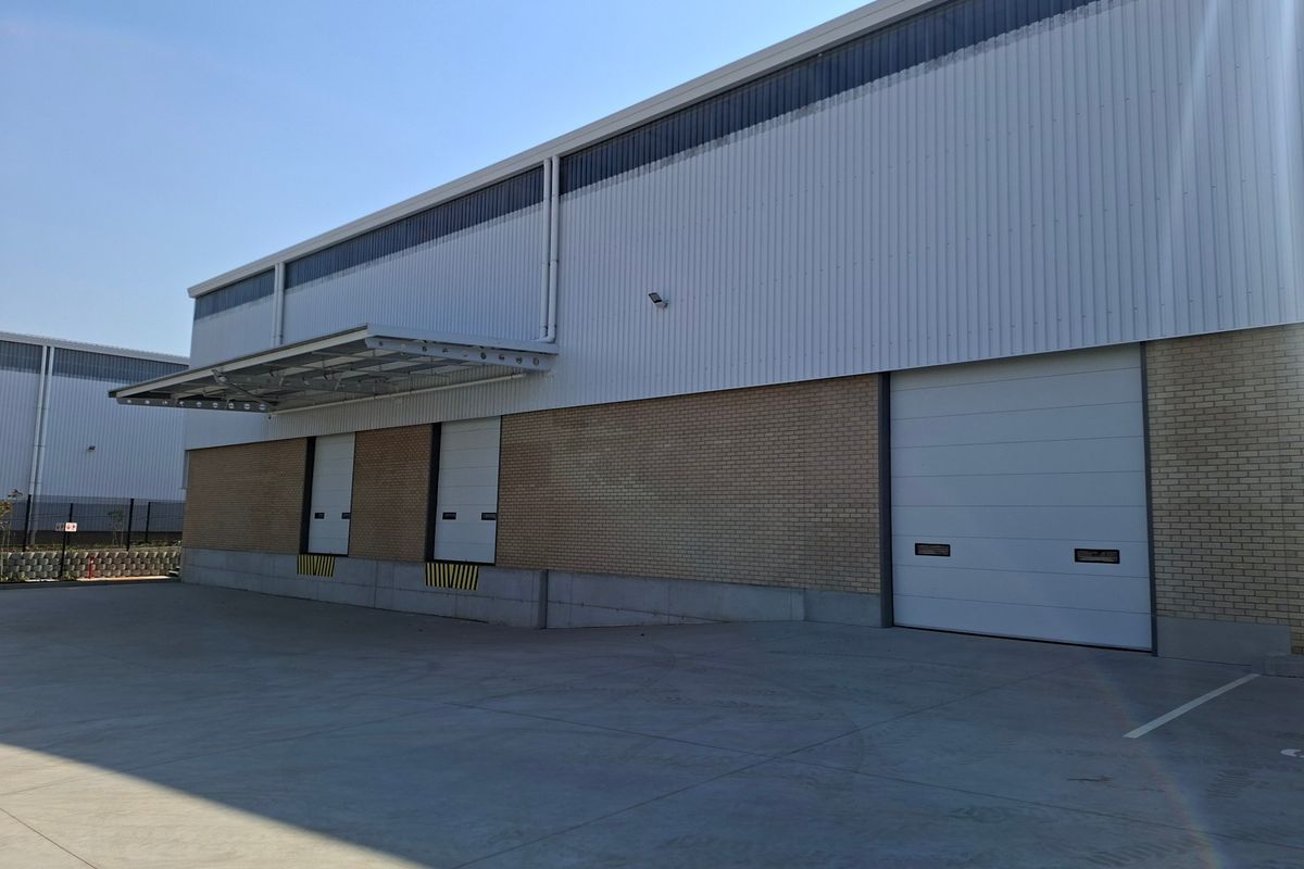Industrial warehouse exterior featuring a concrete loading area, multiple roller shutter doors, and a combination of brick and corrugated metal cladding.