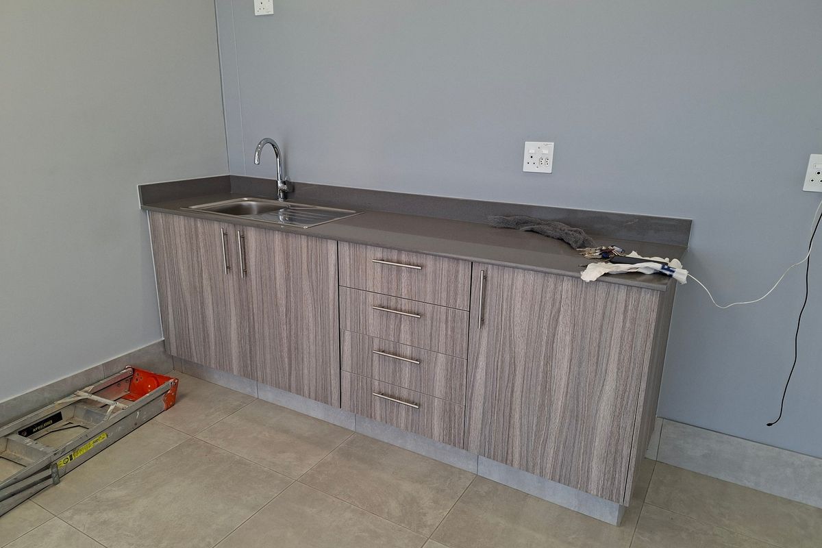 A clean, modern kitchenette area featuring wood-grain cabinetry, a stainless steel sink, and neutral tiled flooring in an industrial space.