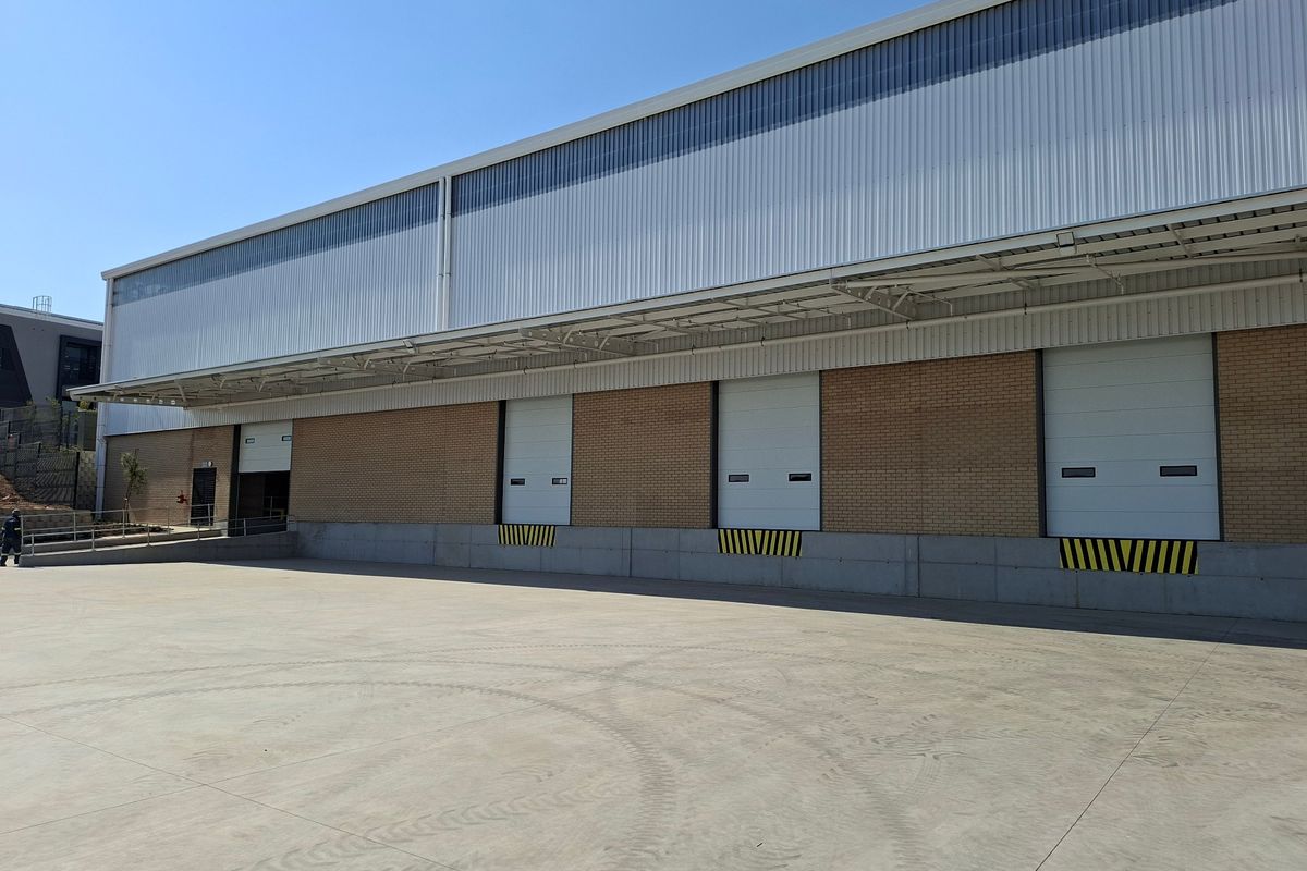 Industrial warehouse exterior featuring multiple roller shutter loading doors, a concrete yard, and modern metal cladding with a protective steel canopy.