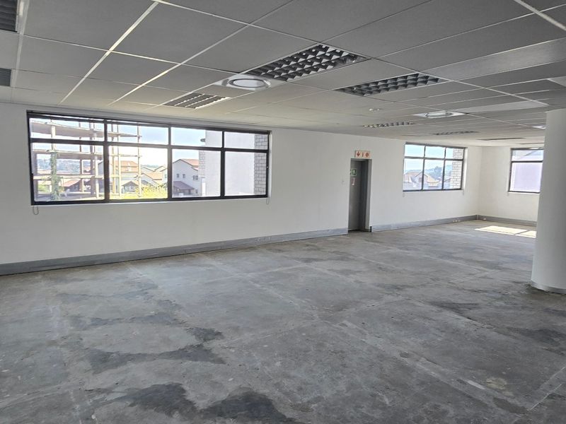 A spacious, vacant commercial office area featuring a suspended ceiling, recessed lighting, large windows providing natural light, and an exposed concrete floor.