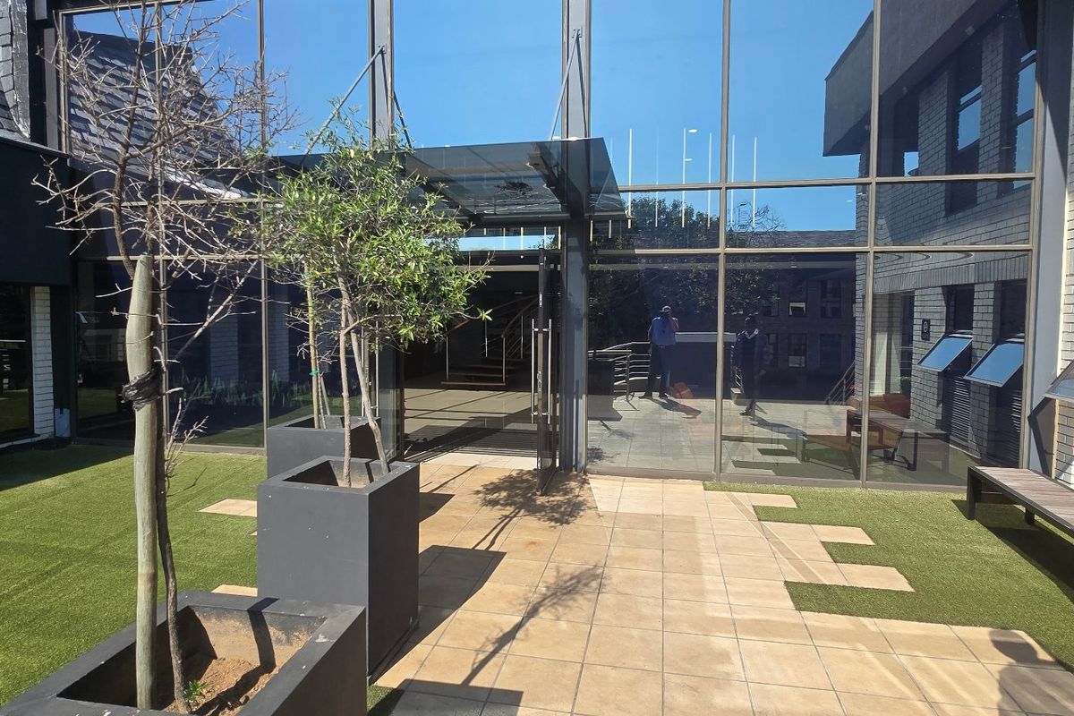 Modern commercial exterior courtyard featuring a glass curtain wall facade, glass entrance canopy, paved patio area, and integrated artificial turf landscaping.