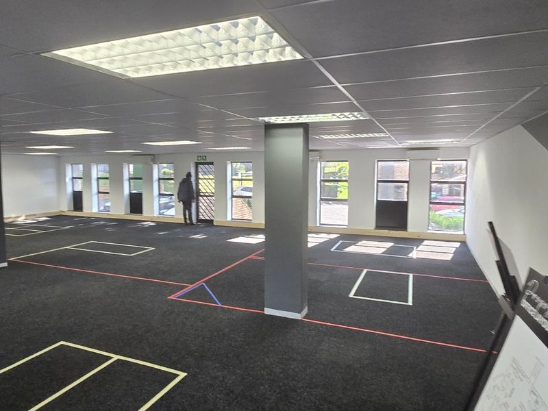 Spacious open-plan commercial office area featuring a suspended ceiling, recessed lighting, structural columns, and large windows providing ample natural light.
