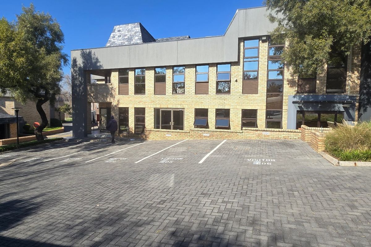 Front exterior view of a commercial office building featuring a brick facade, large aluminium windows, and a paved visitor parking area.