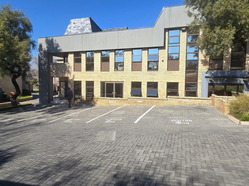 Front exterior view of a commercial office building featuring a brick facade, large aluminium windows, and a paved visitor parking area.