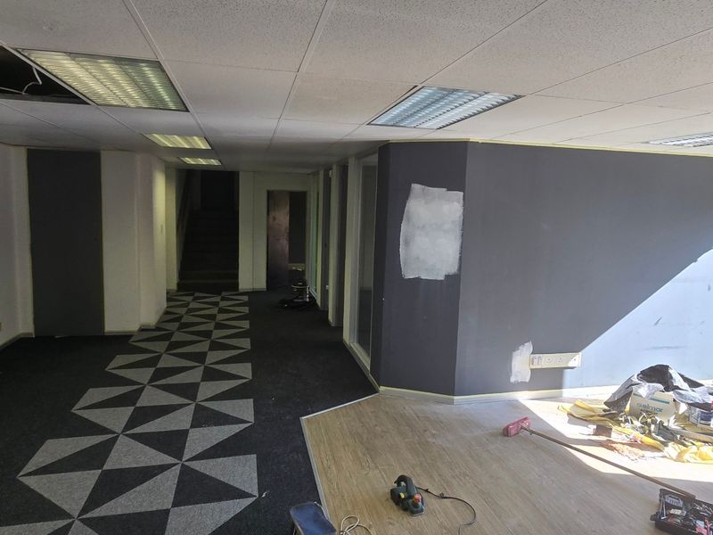 Open-plan commercial office space currently undergoing renovations, featuring a mix of carpet and laminate flooring, suspended ceilings, and large glass entrance doors.