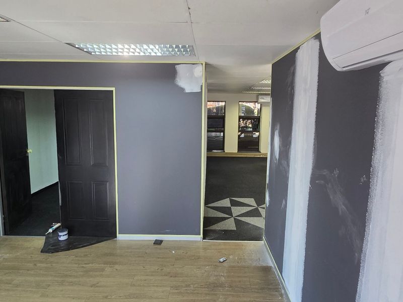 An office space currently undergoing renovation with unfinished wall painting and exposed drywall joints, featuring laminate flooring and a split-system air conditioner.