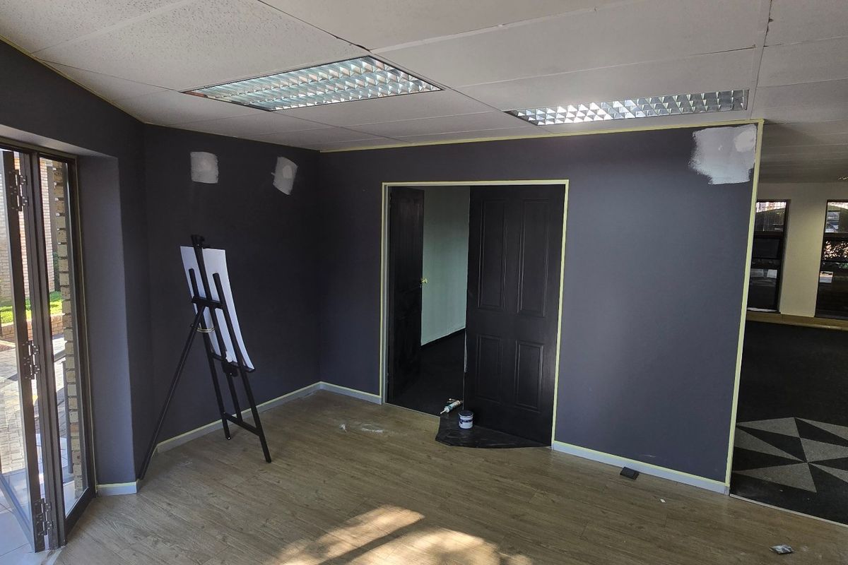 An empty commercial office space featuring laminate flooring, recessed lighting, and a glass entrance door, currently undergoing interior wall finishing and painting.