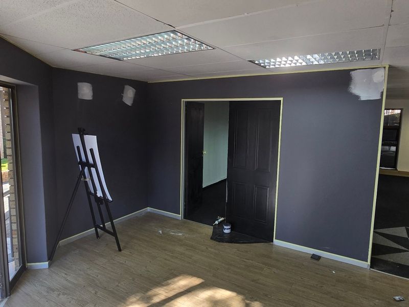 An empty commercial office space featuring laminate flooring, recessed lighting, and a glass entrance door, currently undergoing interior wall finishing and painting.