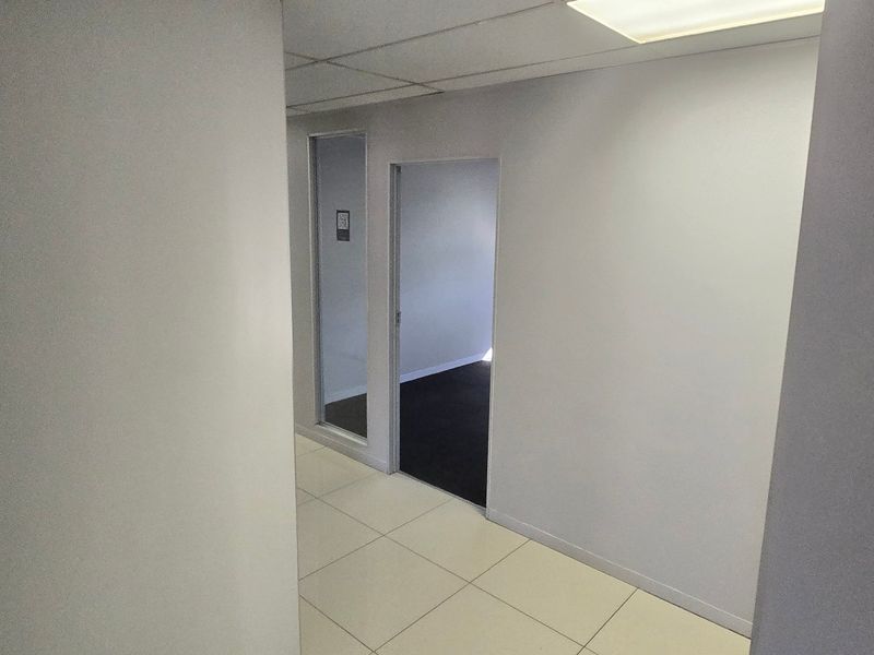 A clean, empty commercial hallway featuring tiled flooring, a suspended ceiling with recessed lighting, and access to an adjacent office space.