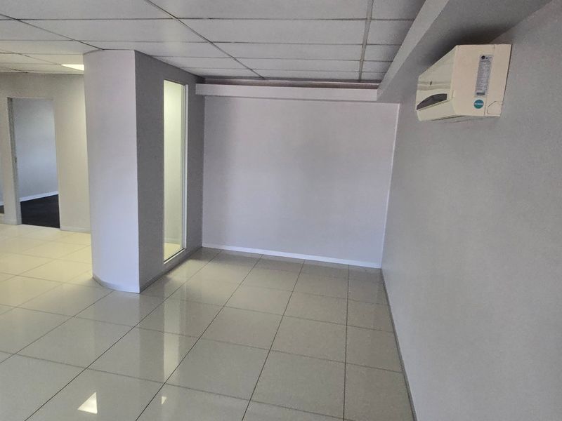 A clean, empty commercial office space featuring tiled flooring, a suspended ceiling, and a wall-mounted air conditioning unit.
