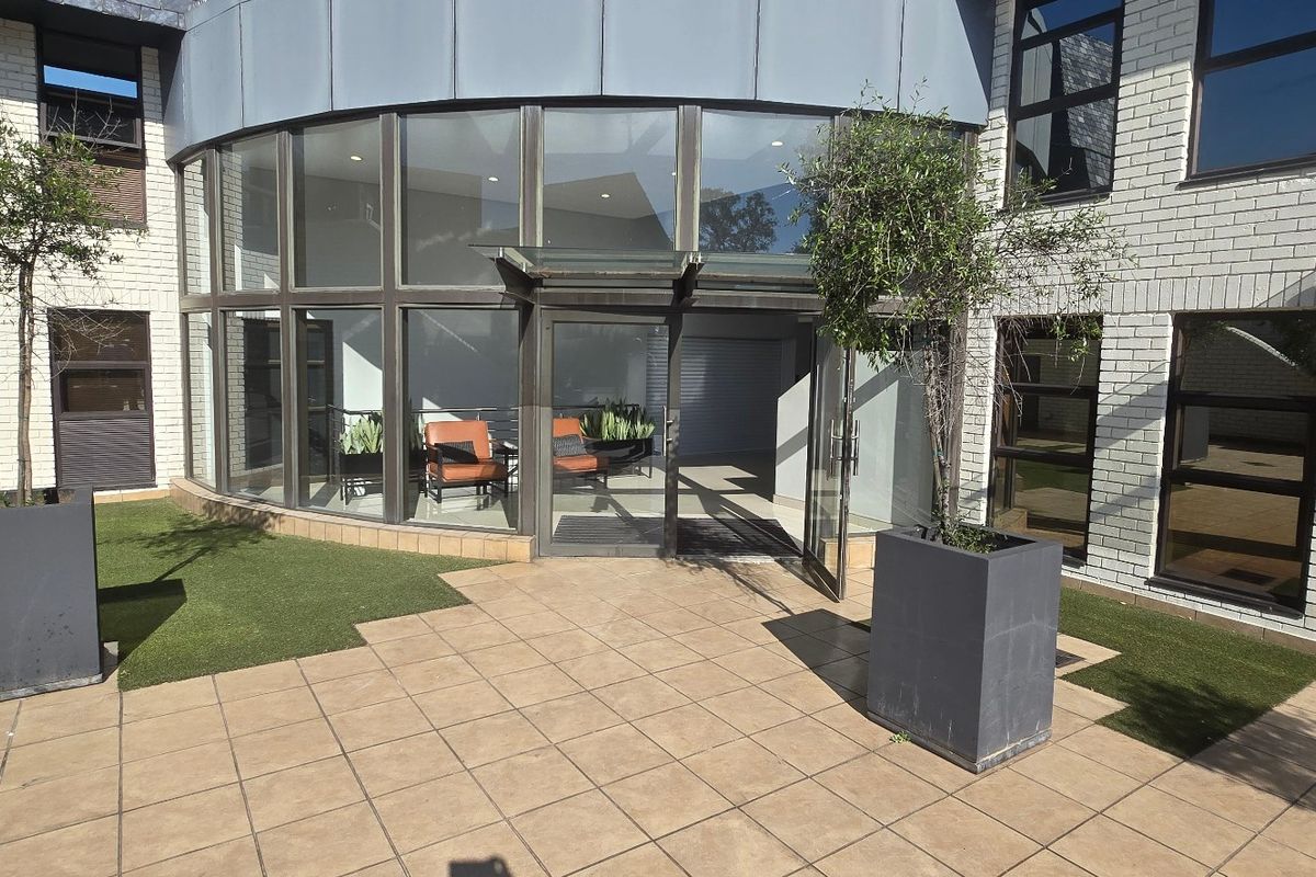 Modern commercial building entrance featuring a curved glass facade, brick exterior, and a tiled patio area with integrated landscaping.