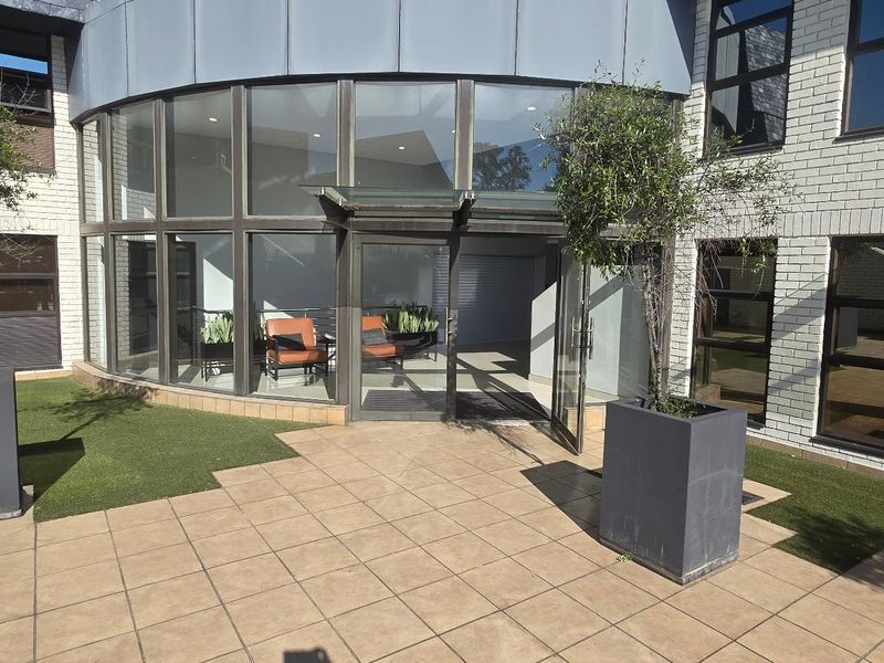 Modern commercial building entrance featuring a curved glass facade, brick exterior, and a tiled patio area with integrated landscaping.