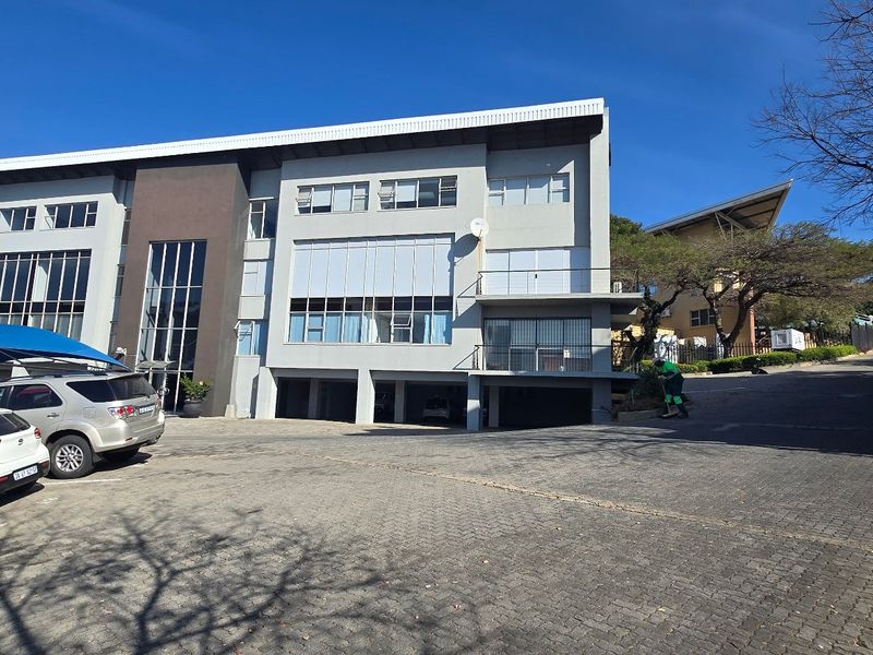 A multi-story commercial office building featuring a paved driveway, undercover parking, and modern aluminium window frames under a clear blue sky.