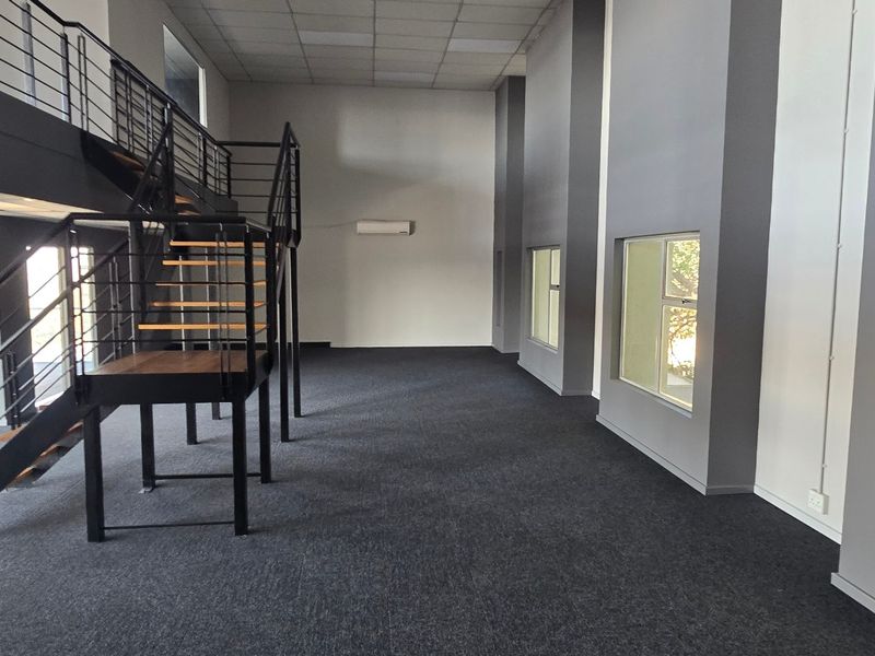 Spacious open-plan office area featuring a modern metal staircase leading to a mezzanine level, large windows, and neutral grey finishes.