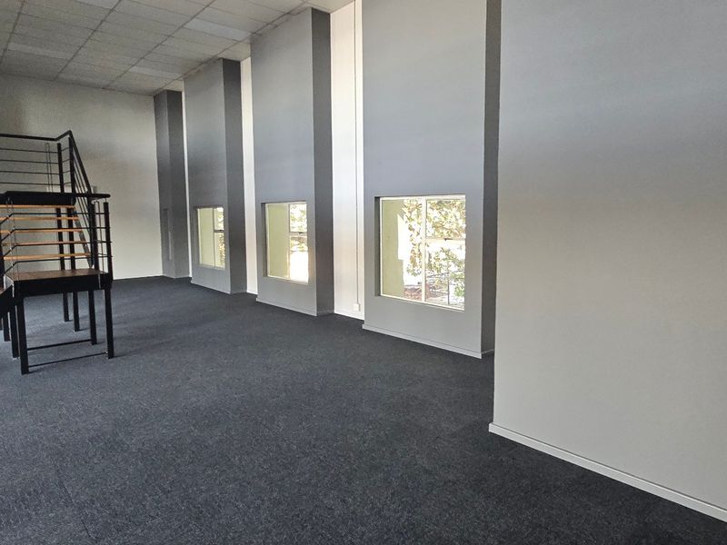 Spacious open-plan commercial office area featuring high ceilings, large windows for natural light, carpeted flooring, and a modern metal staircase.