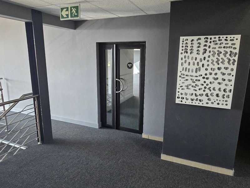 A modern commercial hallway featuring grey carpeting, a glass-panelled entrance door, and a stainless steel staircase railing.