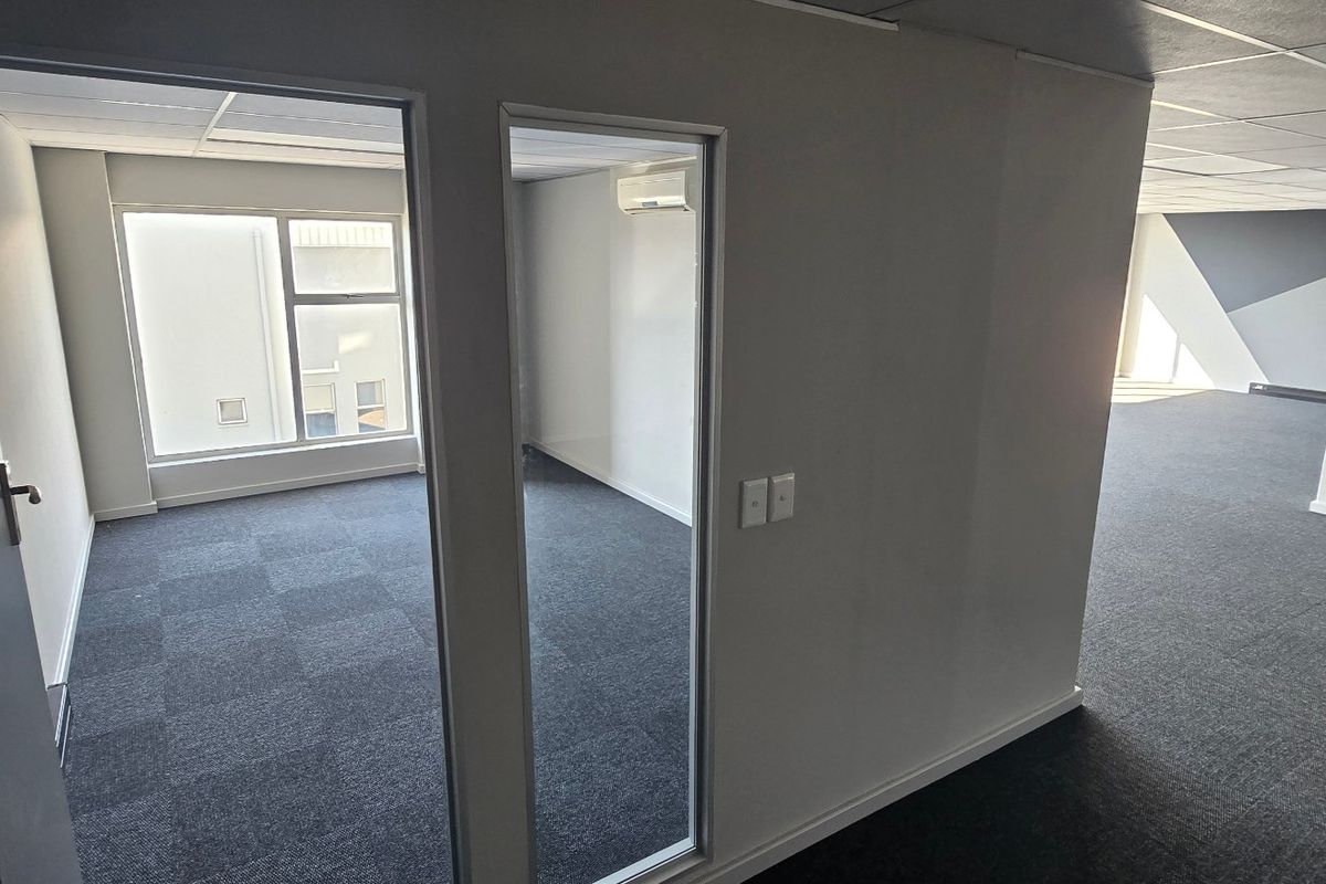 A vacant commercial office space featuring carpeted flooring, a glass-partitioned private office, and a split-system air conditioning unit.