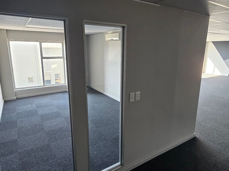 A vacant commercial office space featuring carpeted flooring, a glass-partitioned private office, and a split-system air conditioning unit.