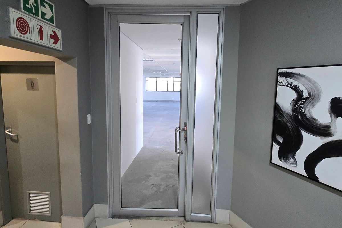 Commercial office entry featuring an aluminium framed glass door, fire safety signage, and neutral grey wall finishes leading into an open plan workspace.