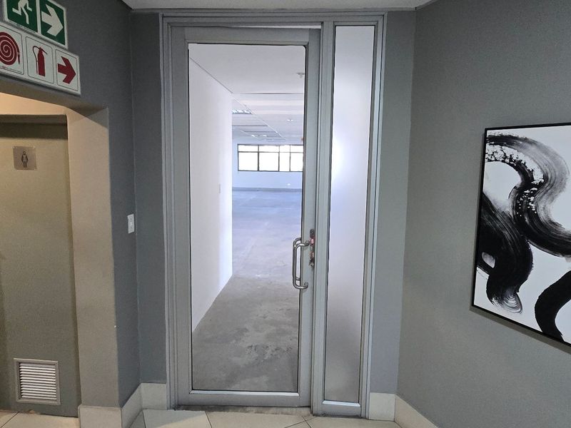Commercial office entry featuring an aluminium framed glass door, fire safety signage, and neutral grey wall finishes leading into an open plan workspace.