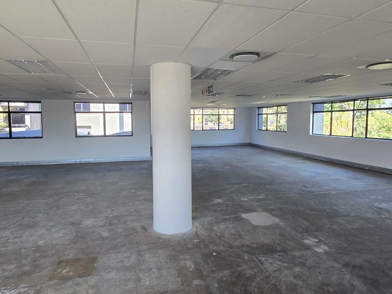 An open-plan commercial office space featuring a suspended ceiling, recessed lighting, a central structural column, and large windows providing natural light.