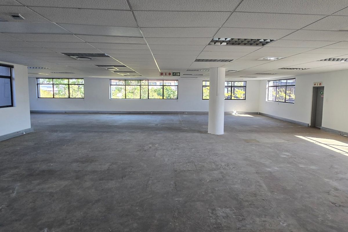 A spacious, open-plan commercial office area featuring a suspended ceiling, structural columns, and large windows providing ample natural light.