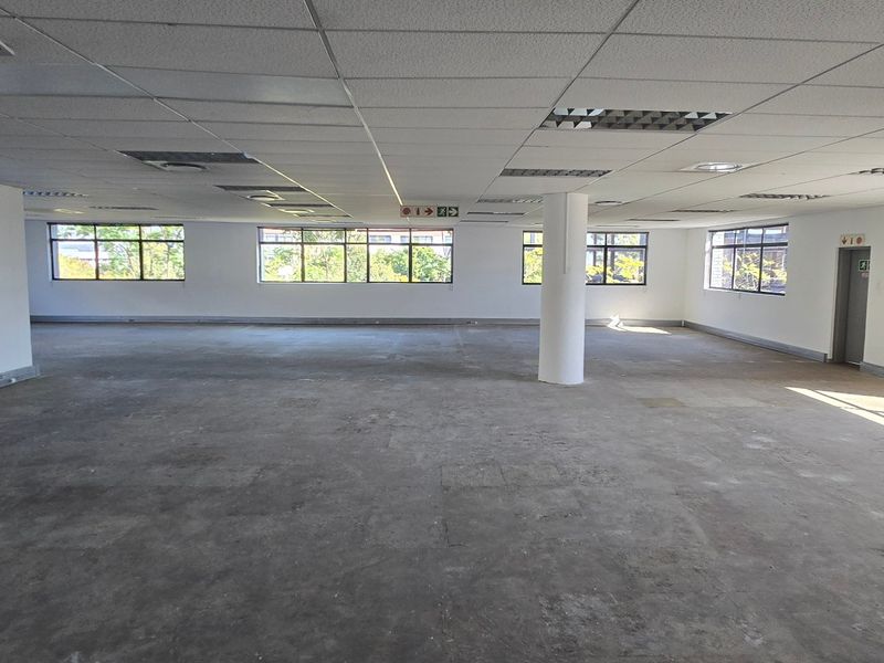 A spacious, open-plan commercial office area featuring a suspended ceiling, structural columns, and large windows providing ample natural light.