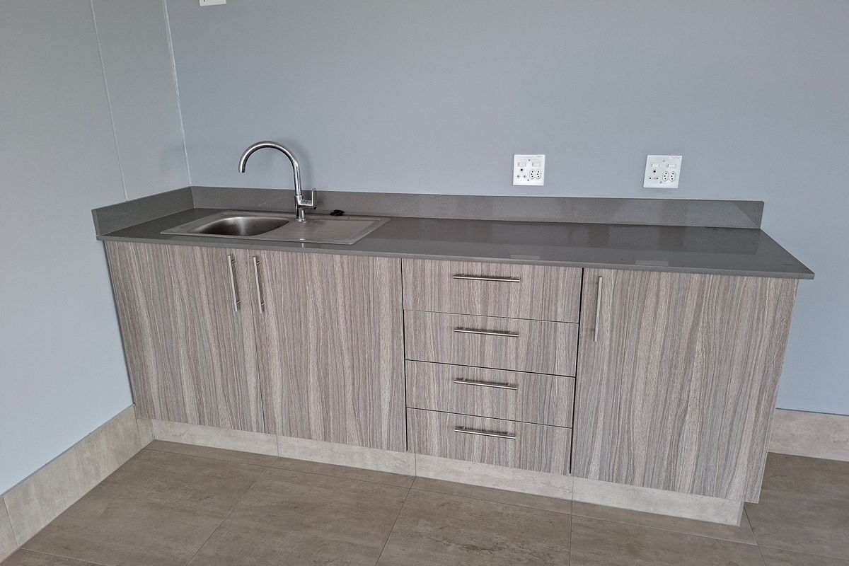 A clean, modern kitchenette featuring wood-grain cabinetry, a stone countertop, and a stainless steel sink, suitable for an office or industrial utility space.