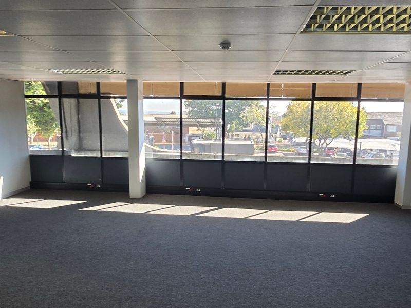 An empty, open-plan office space featuring a large glass window wall, suspended ceiling with recessed lighting, and carpeted flooring.
