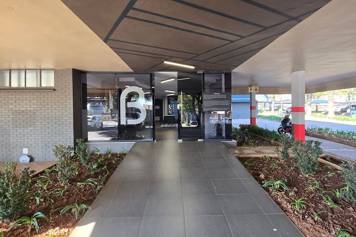 Modern commercial building entrance featuring a covered walkway, glass doors, and landscaped garden beds leading to the main reception area.