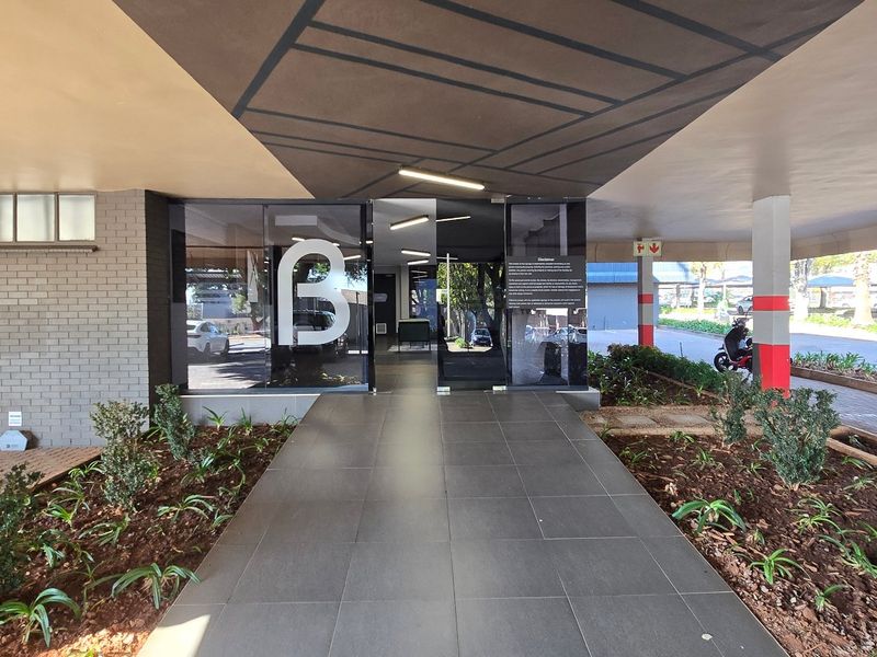 Modern commercial building entrance featuring a covered walkway, glass doors, and landscaped garden beds leading to the main reception area.