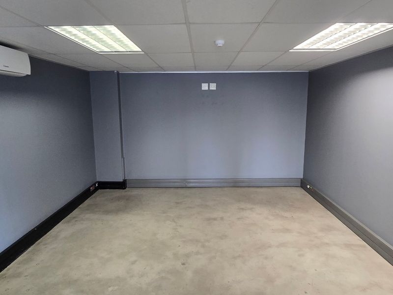 A vacant commercial office space featuring a suspended ceiling, recessed lighting, and a wall-mounted air conditioning unit with grey painted walls.