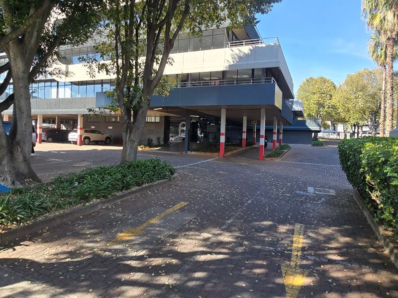Exterior view of a multi-story commercial office building featuring a paved driveway, covered parking area, and established landscaping.