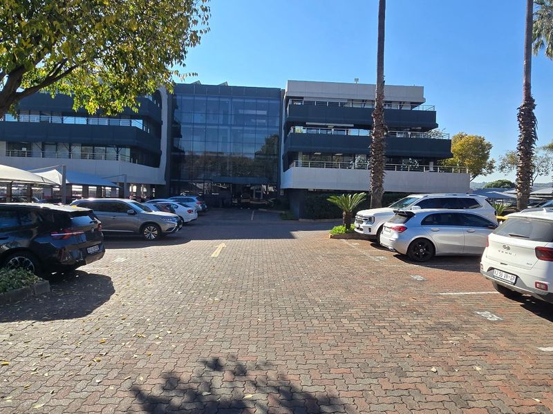 Front view of a multi-story commercial office building featuring a glass facade and an extensive paved parking area with covered bays.