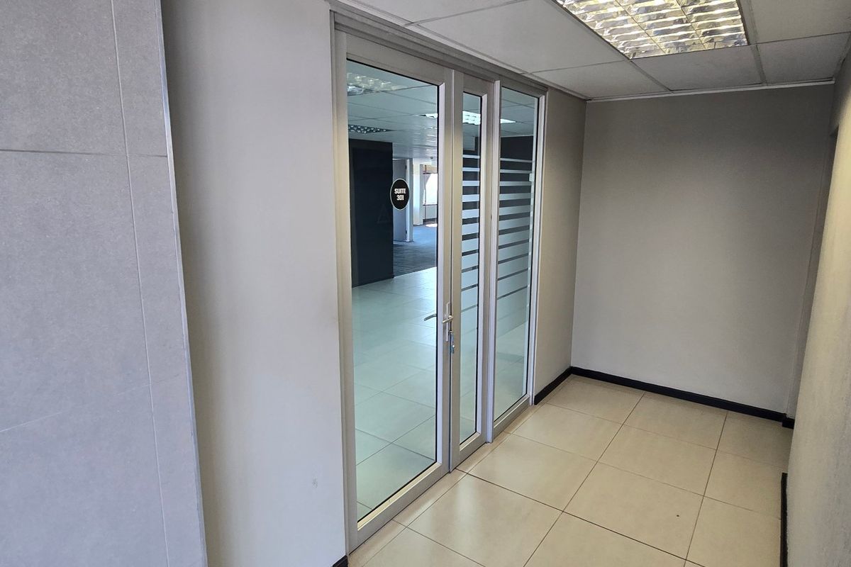 A clean, modern commercial hallway featuring tiled flooring, a suspended ceiling with recessed lighting, and an aluminium-framed glass entrance door.