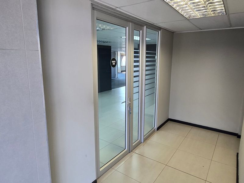 A clean, modern commercial hallway featuring tiled flooring, a suspended ceiling with recessed lighting, and an aluminium-framed glass entrance door.