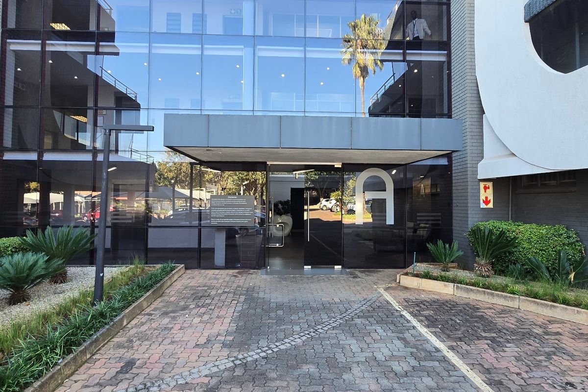 Modern commercial office building entrance featuring a glass curtain wall facade, paved driveway, and well-maintained landscaped garden beds.