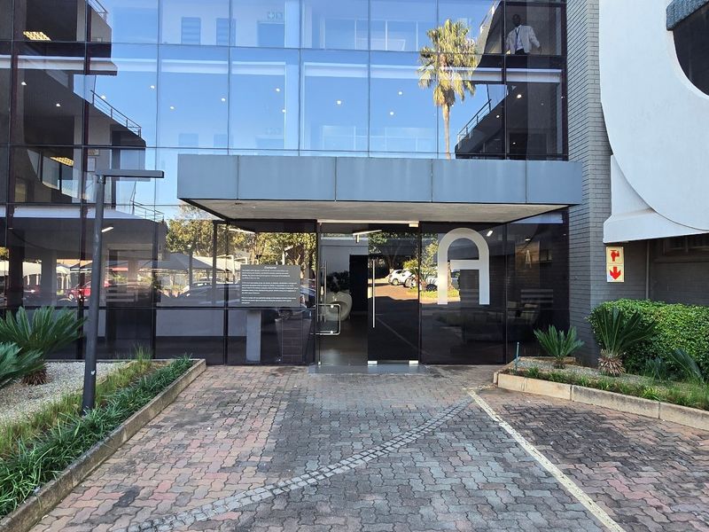 Modern commercial office building entrance featuring a glass curtain wall facade, paved driveway, and well-maintained landscaped garden beds.