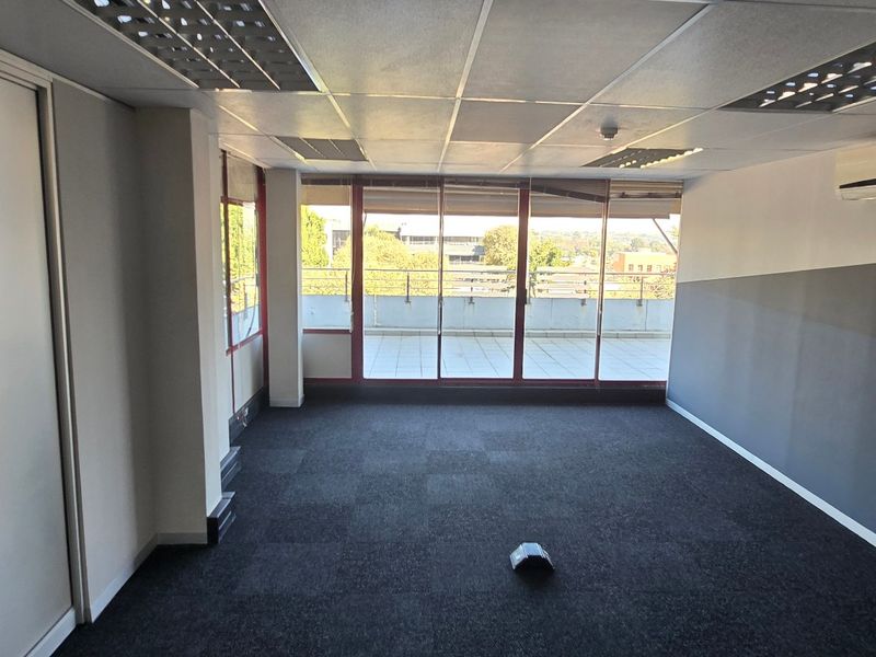 A bright, empty office space featuring carpet tiles, a suspended ceiling with recessed lighting, a wall-mounted air conditioner, and access to a balcony.