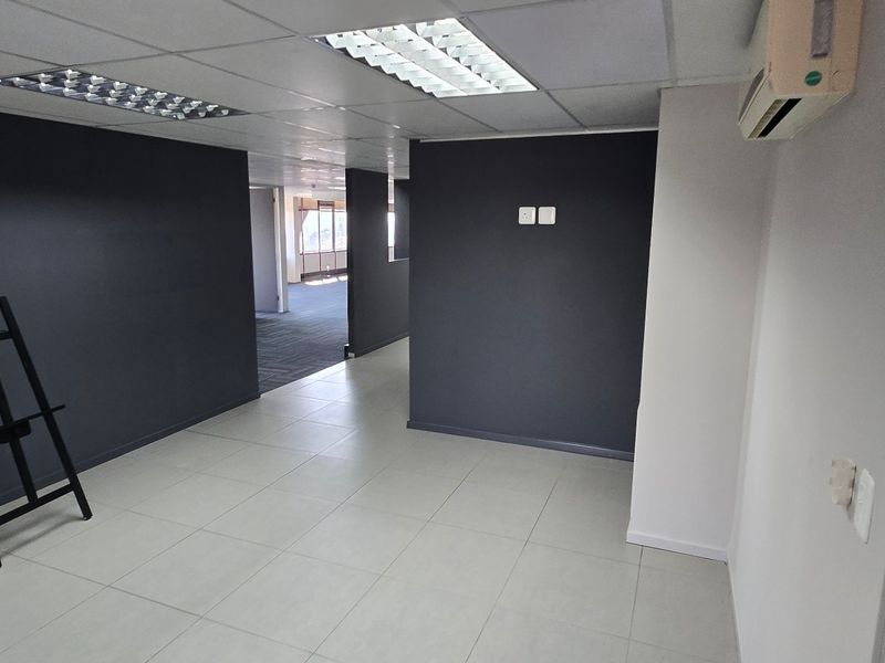 A vacant commercial office space featuring tiled flooring, a suspended ceiling with recessed lighting, and a wall-mounted air conditioning unit.